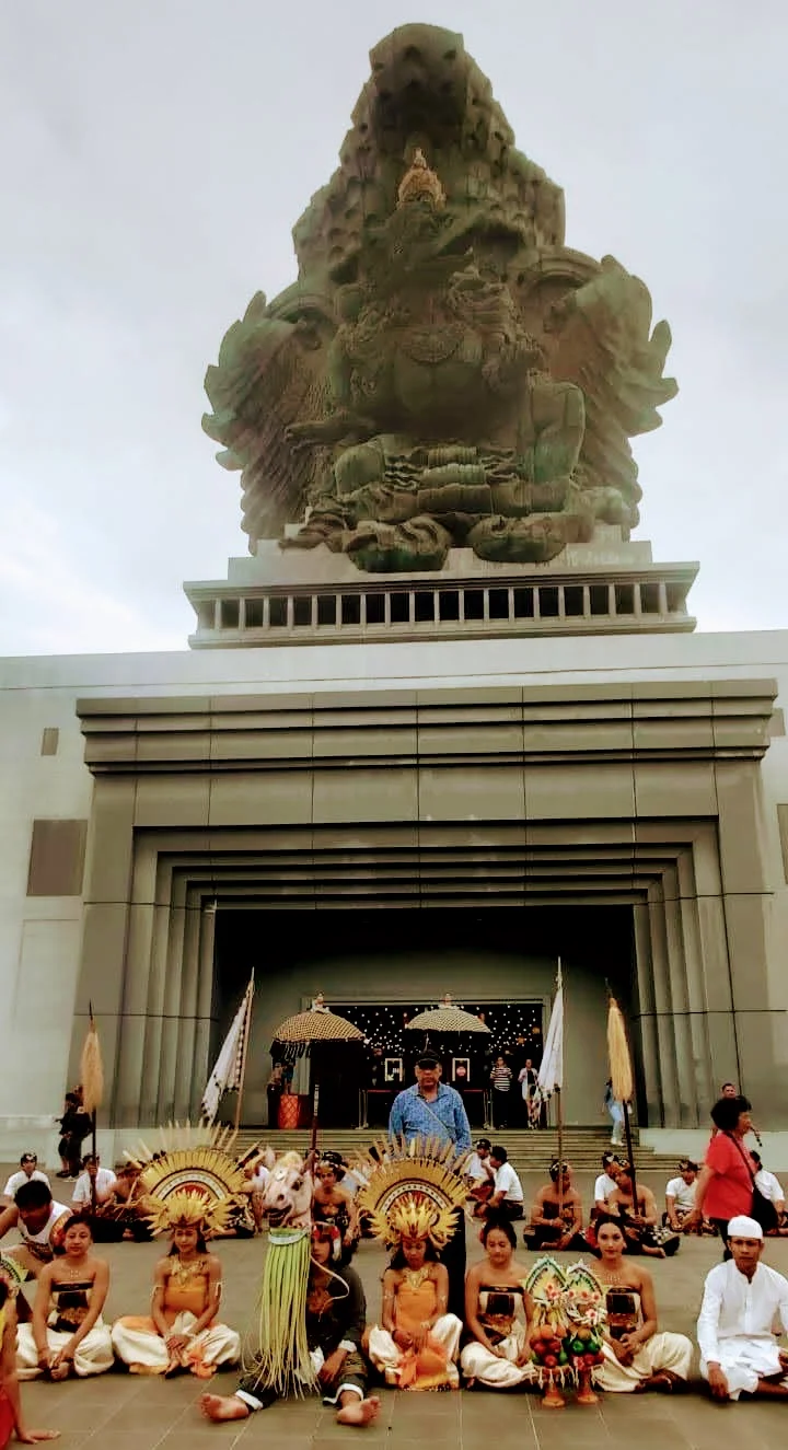 Traveler standing with the full Balinese dance troupe with GWK monument towering above