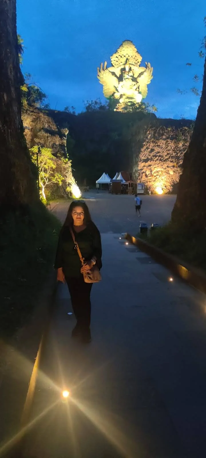 Traveler on the path beneath the illuminated colossal Garuda Wisnu Kencana monument glowing at night in Bali