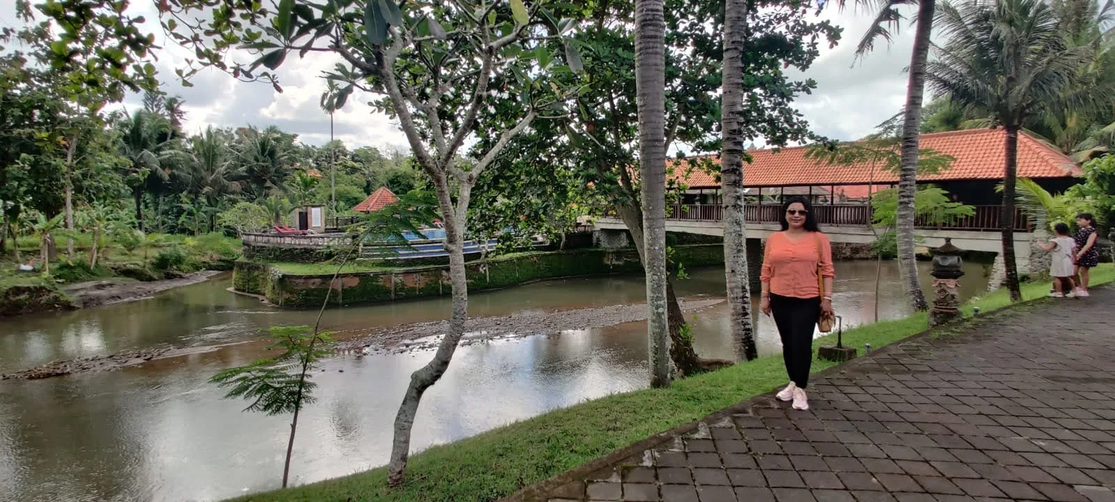 Traveler by the resort riverside promenade with palm trees, tropical flora, and covered footbridge