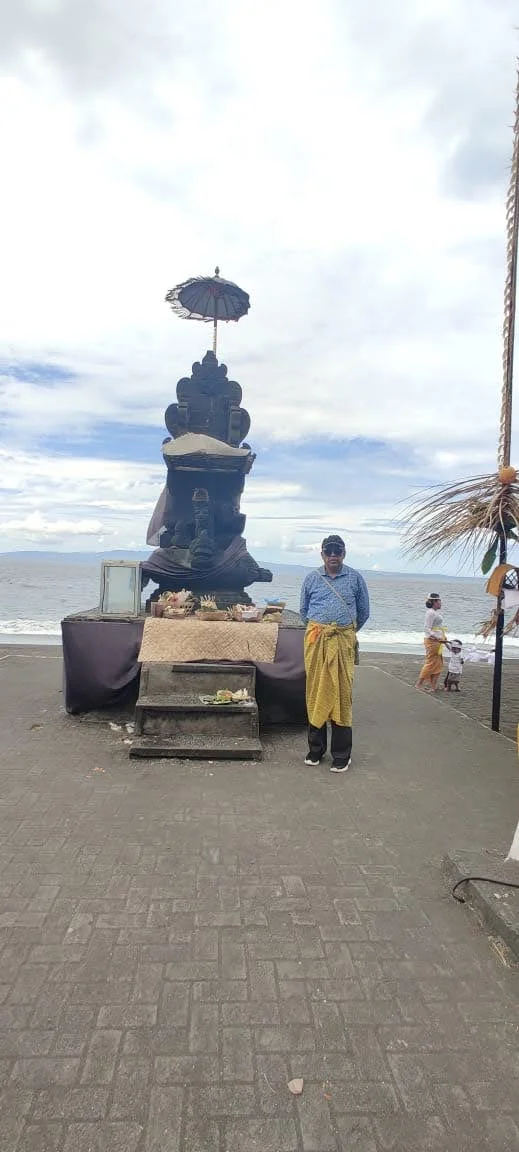 Gentleman traveler in sarong standing beside a seaside padmasana stone altar with offerings and ocean horizon
