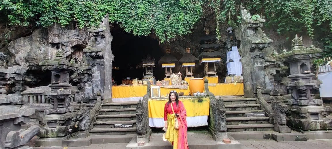 Lady traveler standing before the sacred cave entrance and shrines of Pura Goa Lawah draped in ceremonial yellow