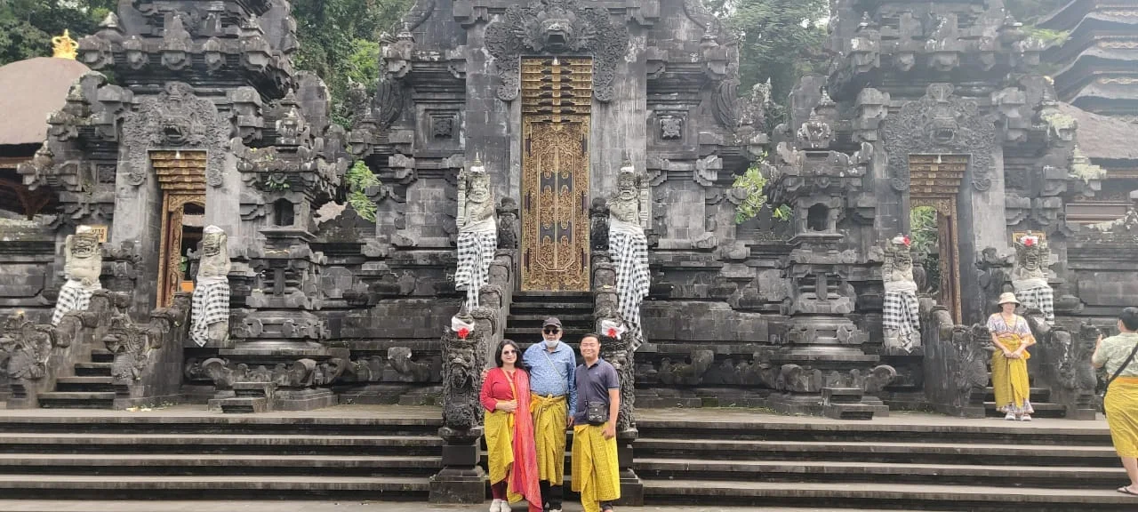 Panoramic view of both travelers and their guide before the three monumental Kori Agung stone portals at Goa Lawah