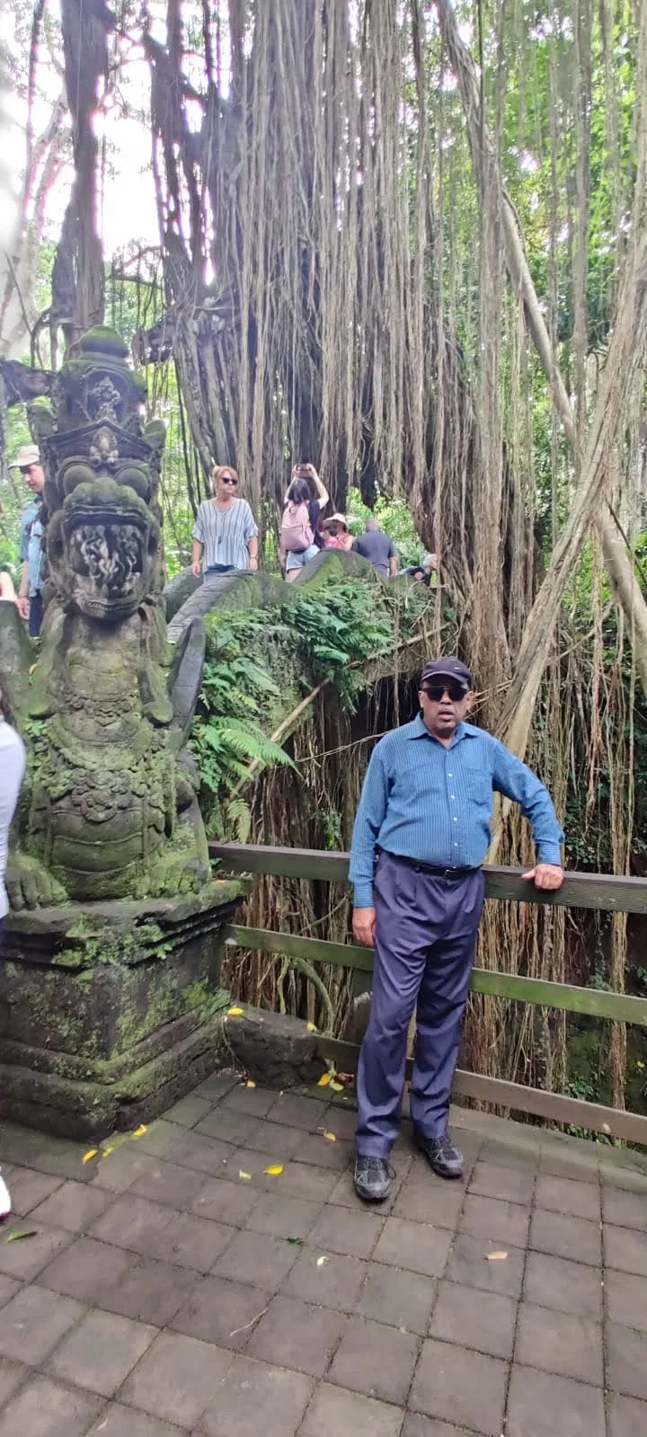 Gentleman traveler beside a moss-covered dragon statue beneath colossal banyan aerial roots in Ubud Monkey Forest