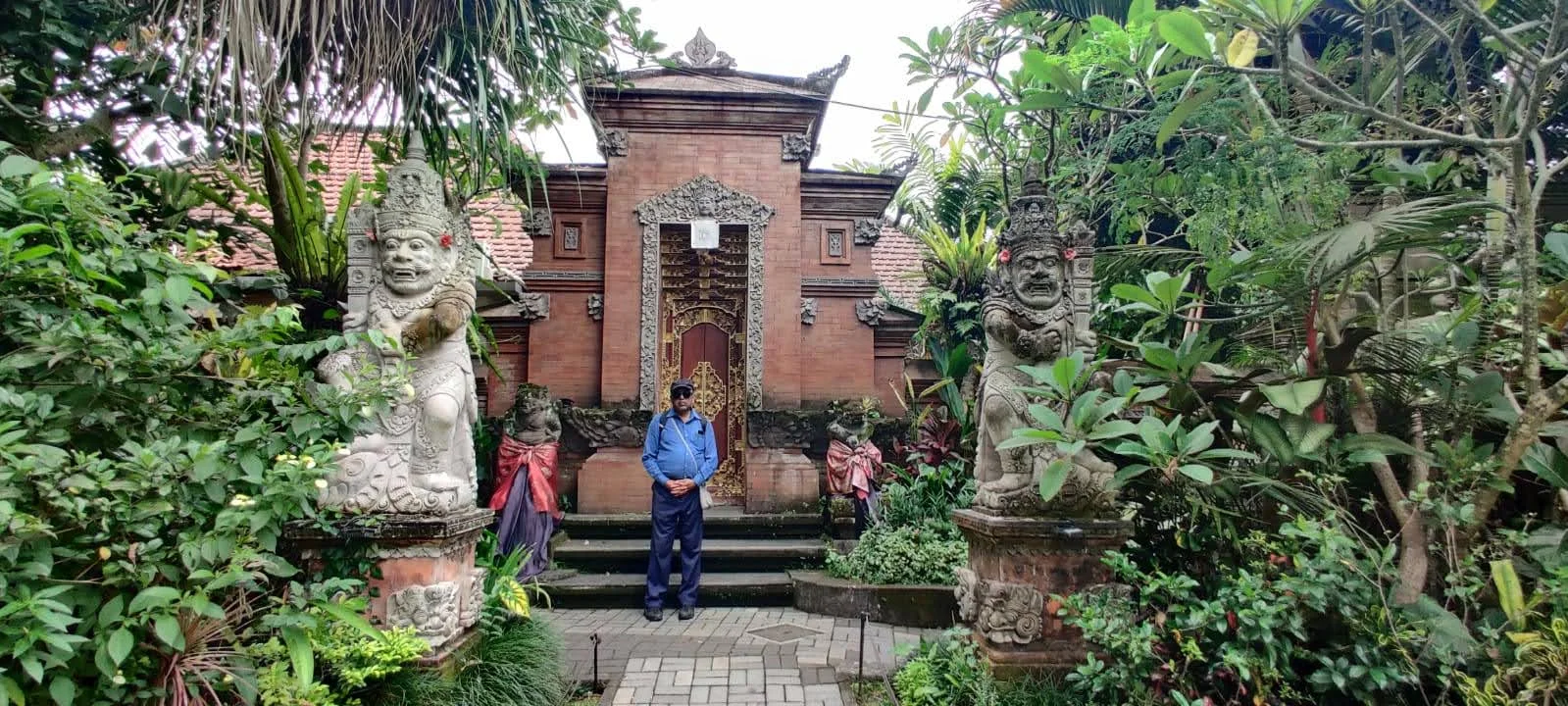 Gentleman traveler between twin carved stone guardian statues adorned with red hibiscus flowers in Ubud Palace garden