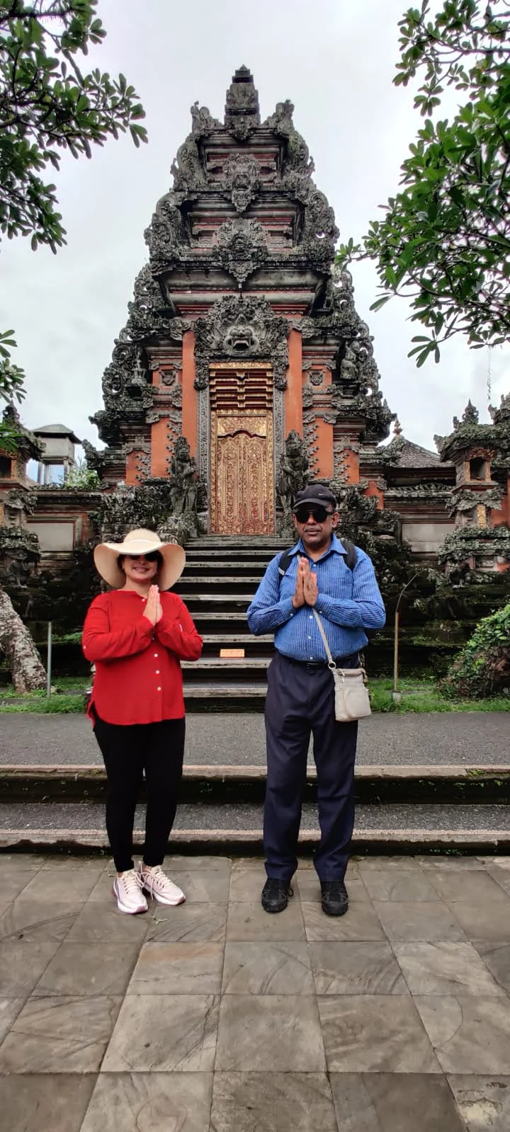 Both travelers in Namaste prayer pose before the towering carved stone portal and gilded doors of Pura Taman Saraswati