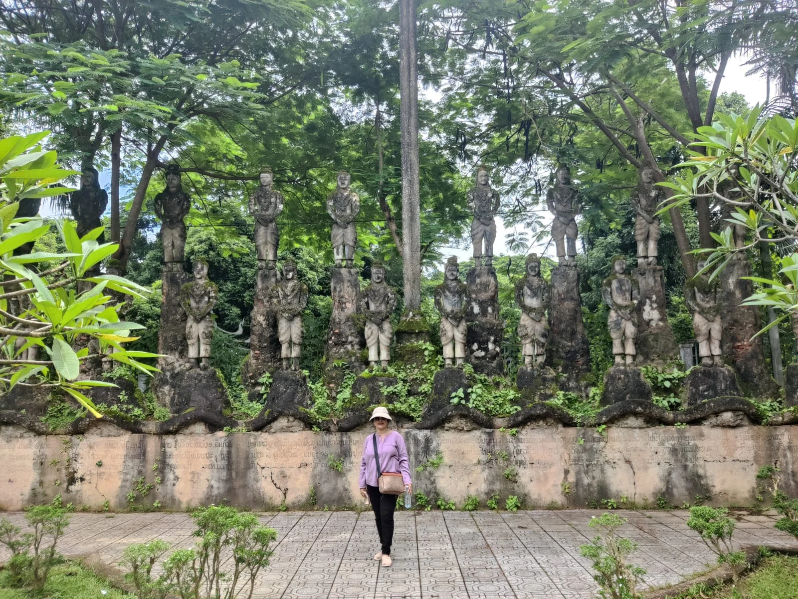 Traveler beside a long curved balustrade wall lined with dozens of multi-armed concrete guardian deities and celestial devas
