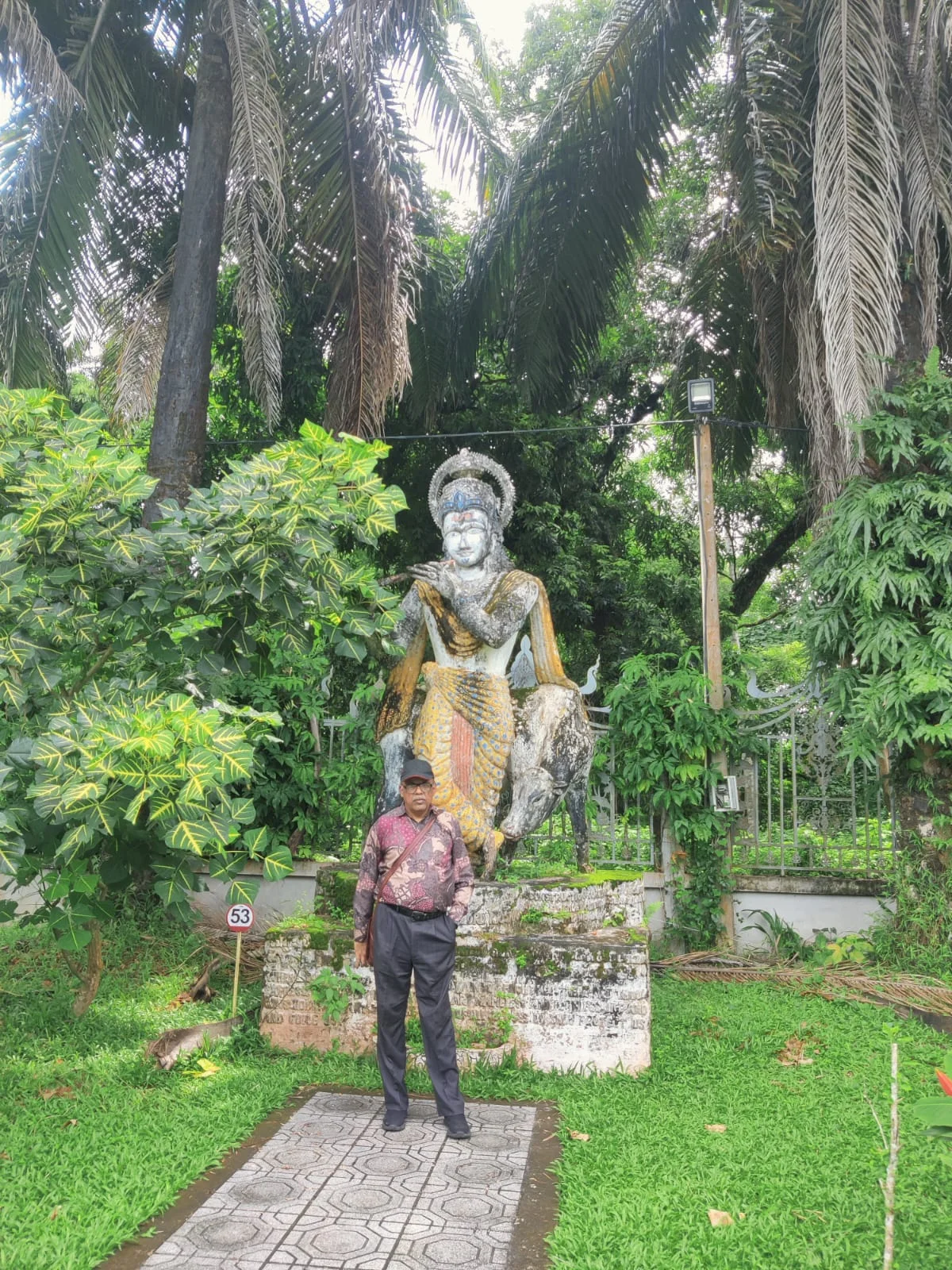 Traveler posing beside the detailed concrete sculpture of Lord Krishna playing the flute accompanied by a cow and multi-armed deities