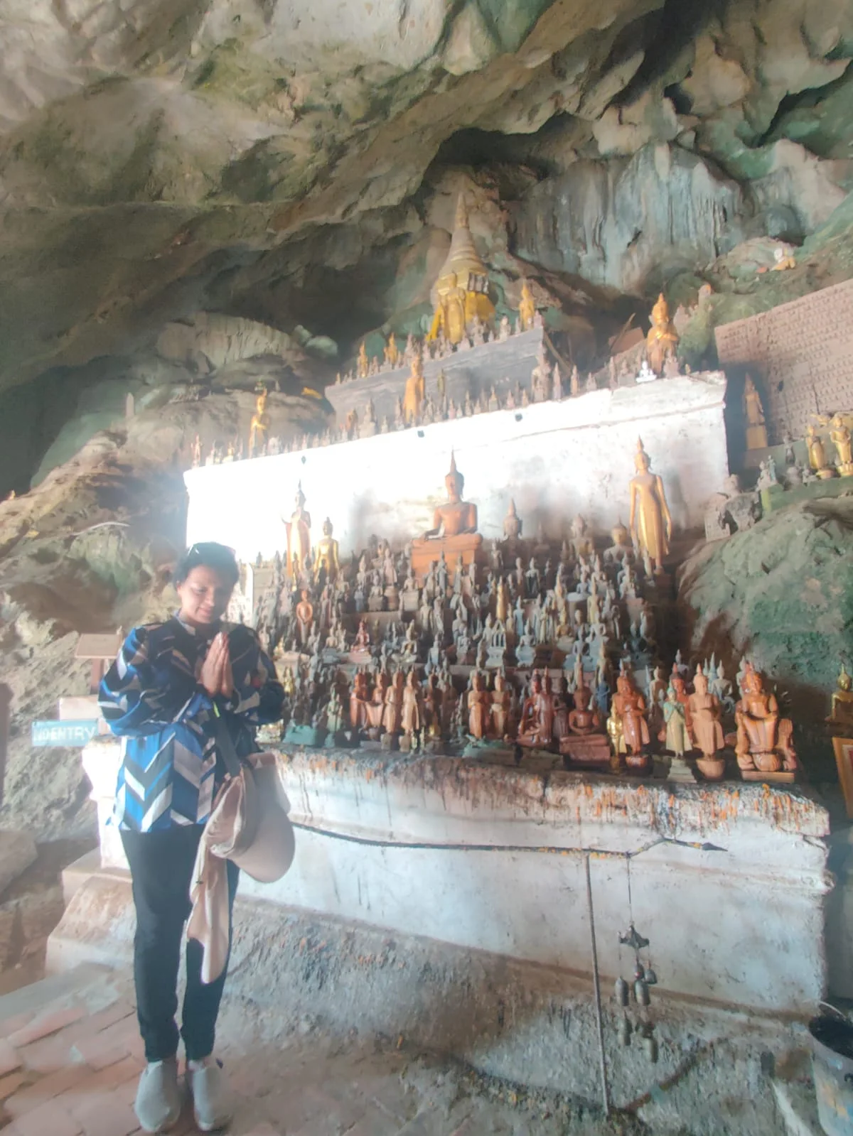 Female traveler standing reverently before ancient limestone ledge shrines crowded with miniature wooden and bronze Buddha statues in Pak Ou Caves