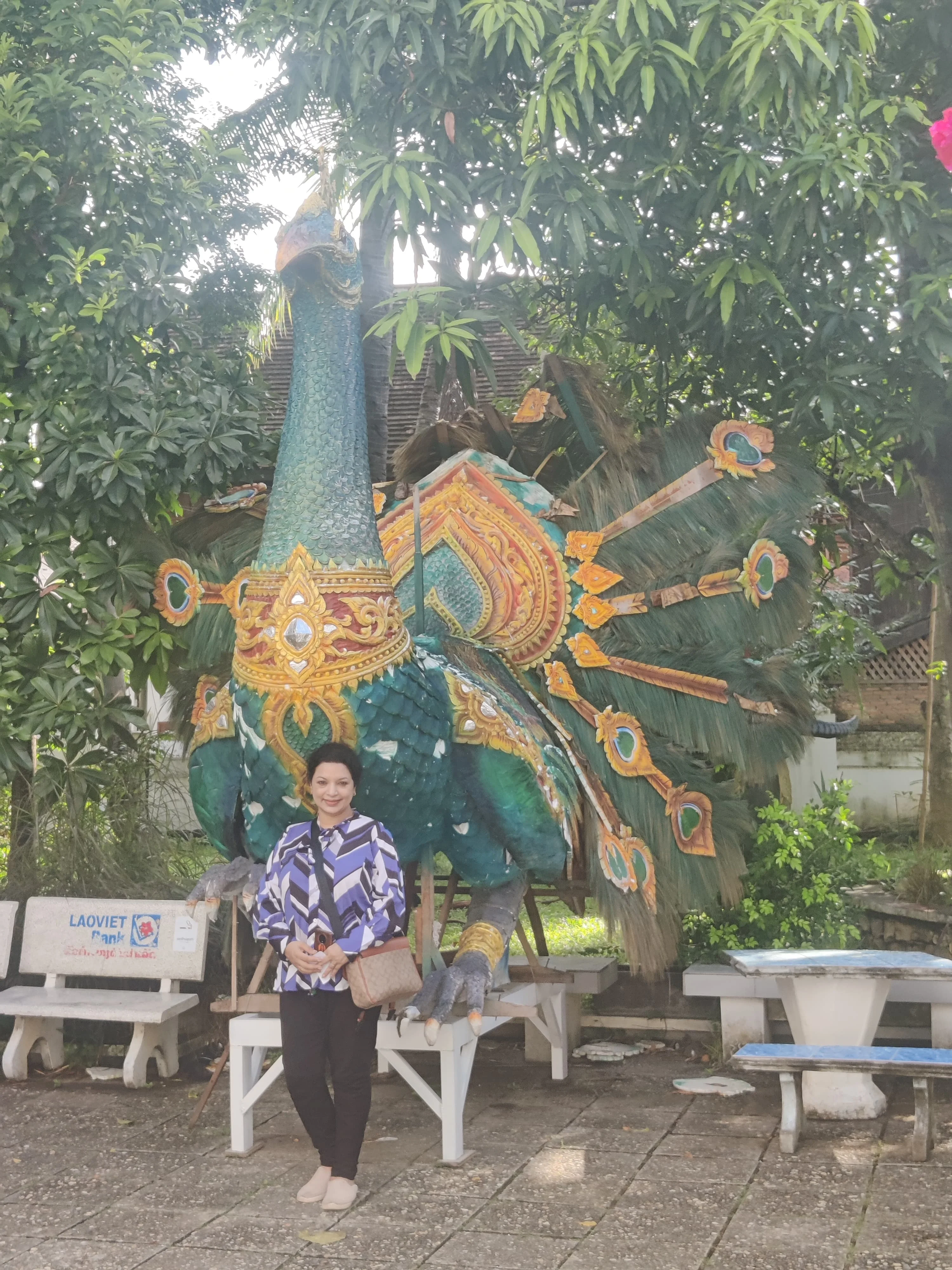 Female traveler beside an ornate carved golden Mayura peacock guardian sculpture in the landscaped gardens of Wat Xieng Thong