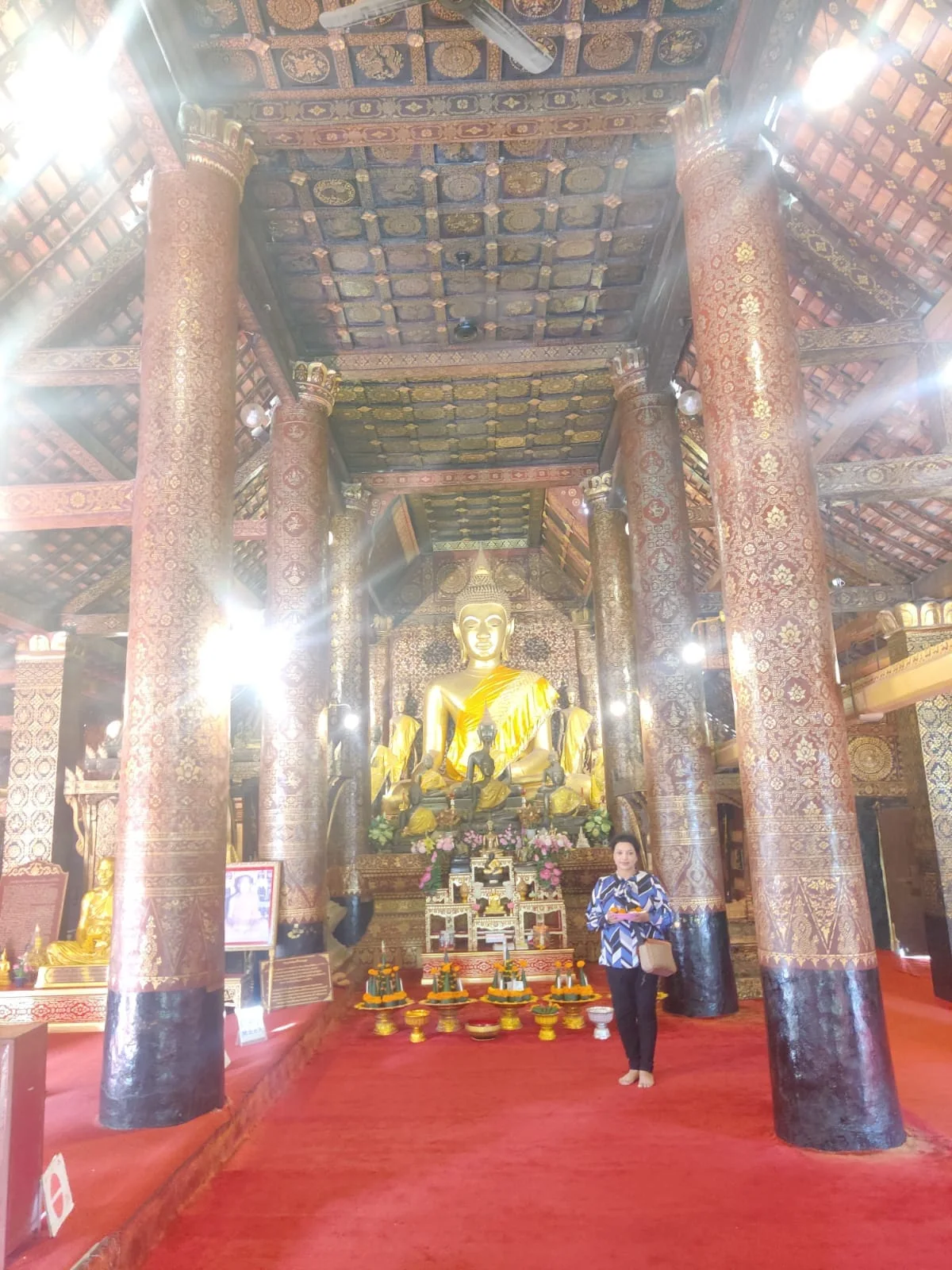 Female traveler inside the dim, serene sanctuary of Wat Xieng Thong surrounded by gold-stenciled lacquer pillars before the gilded Buddha