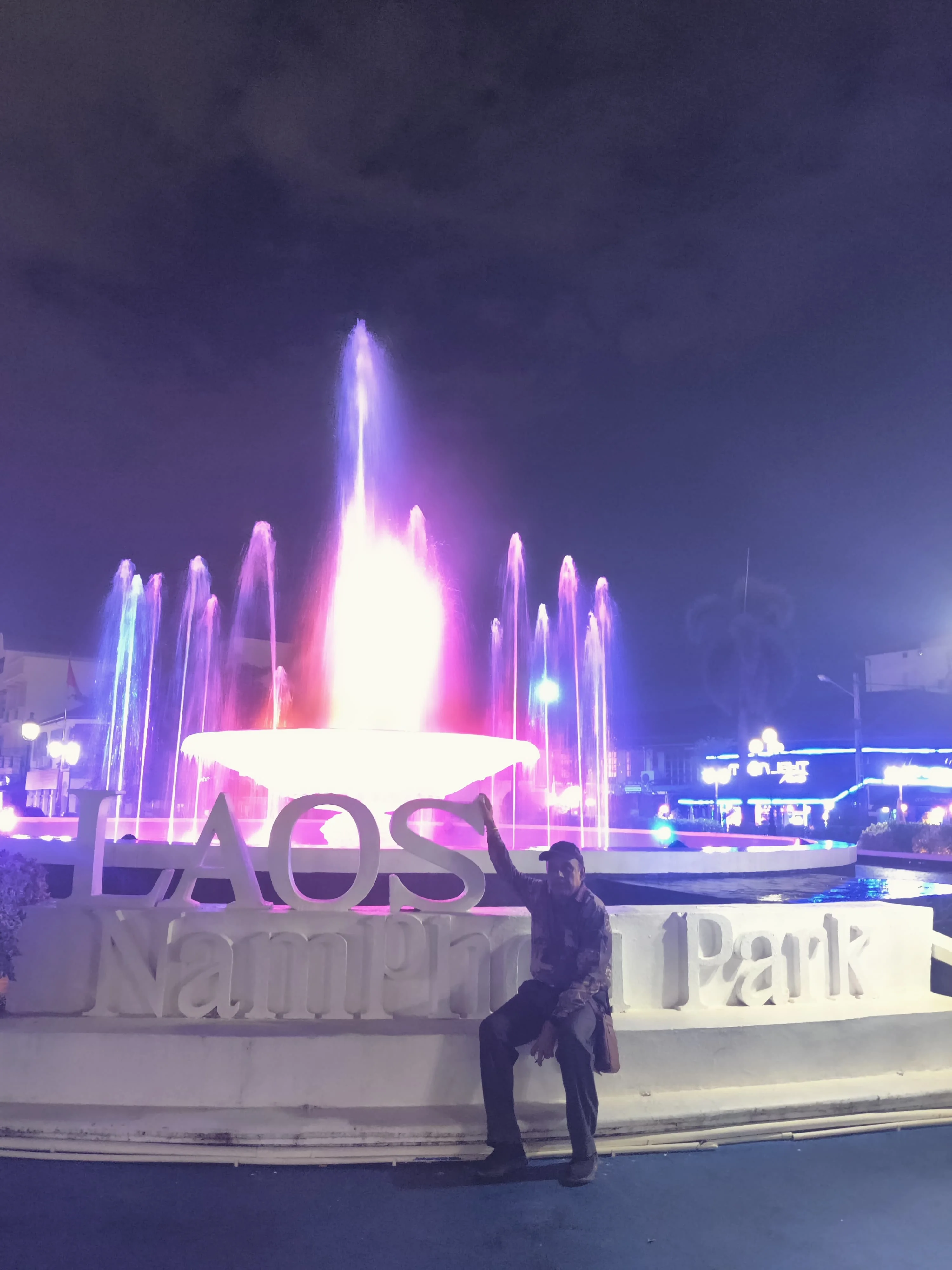 Male traveler in Nam Phou Fountain square in central Vientiane illuminated with colorful fountain jets and evening bistro lights