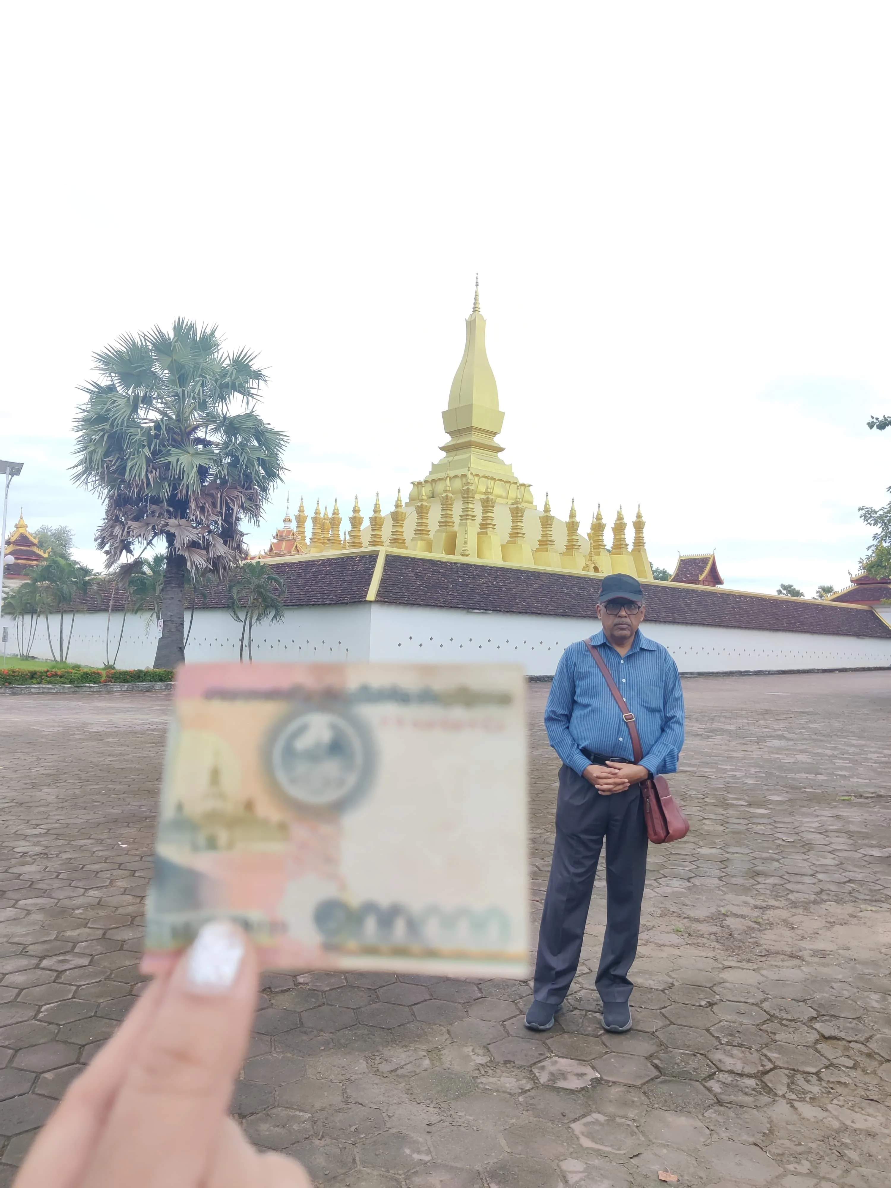 Male traveler holding up a Lao Kip banknote showing the Pha That Luang stupa aligned with the real golden monument