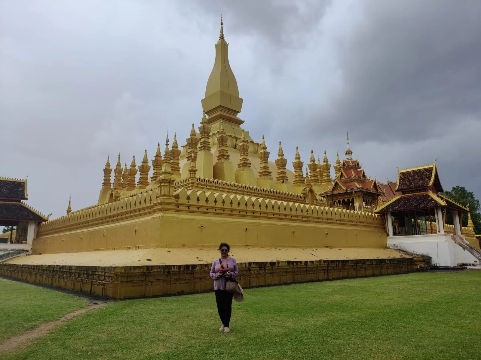 Female traveler smiling before the colossal shimmering golden spire of Pha That Luang under brilliant skies in Vientiane