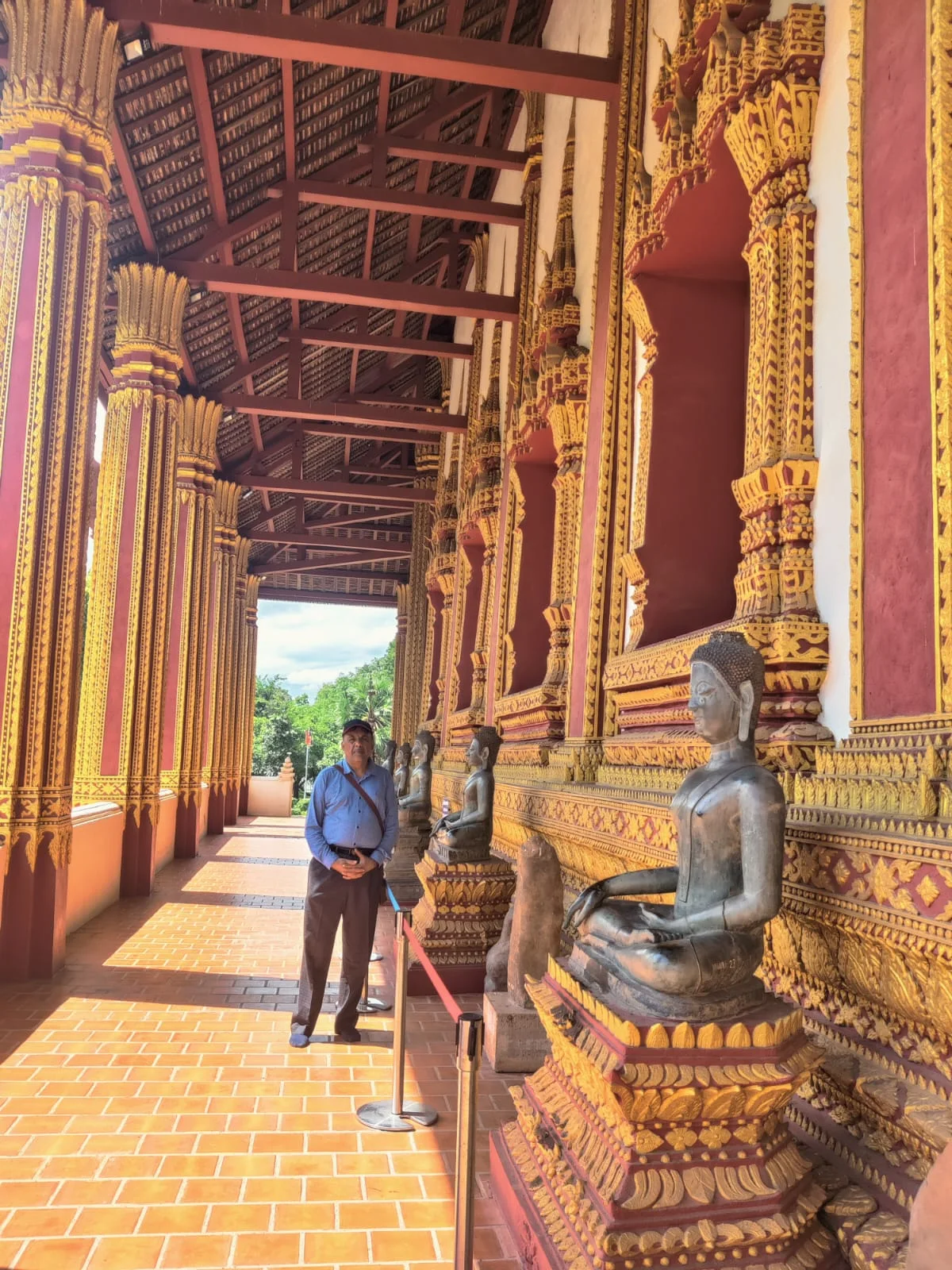 Male traveler walking along the open-air peristyle gallery of Wat Ho Phra Keo beside ancient bronze Buddha statues displaying historic Lao mudras