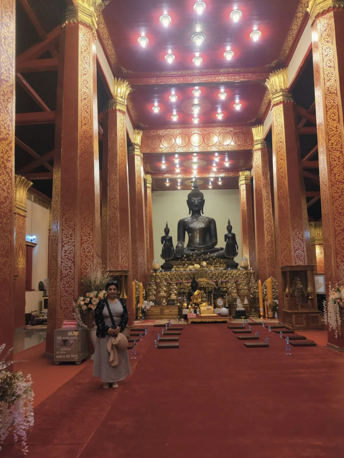 Female traveler seated with folded hands in the sanctum of Wat Ong Teu before the monumental bronze seated Buddha flanked by red and gold lacquered pillars