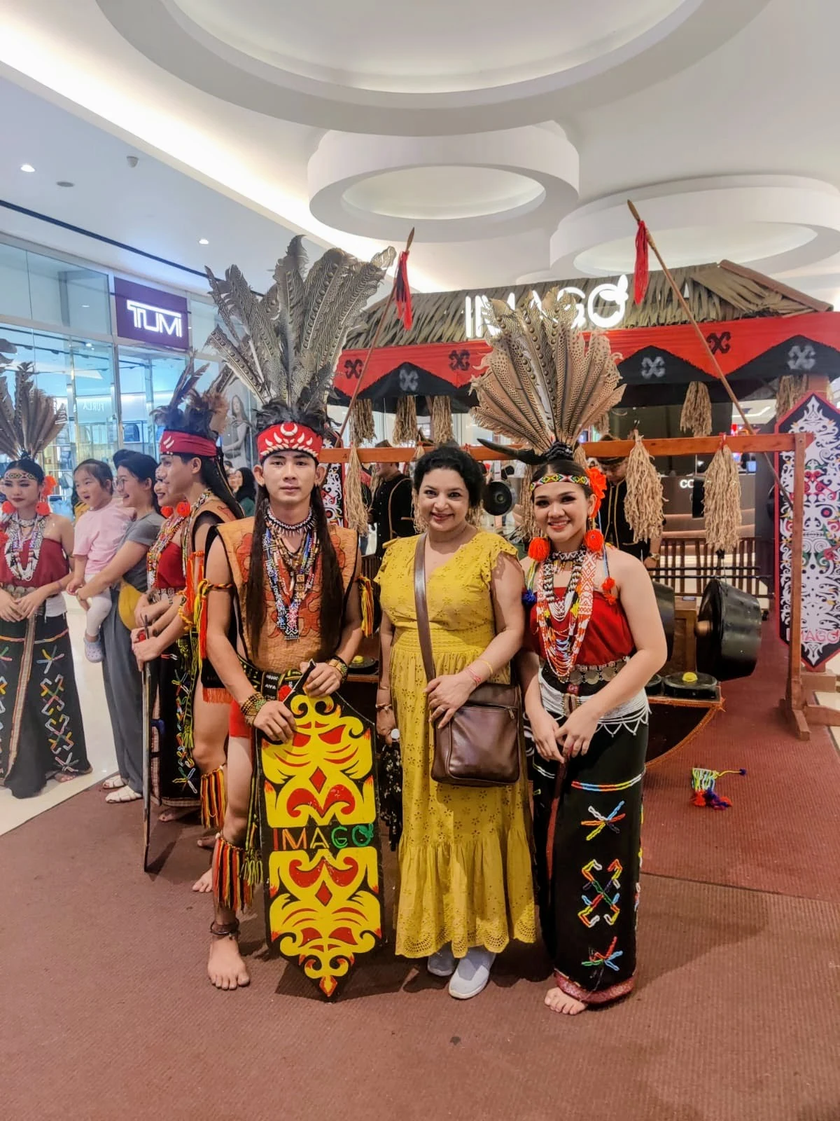 Female traveler smiling alongside Dayak and Murut indigenous performers in elaborate feather headdresses and traditional woven regalia