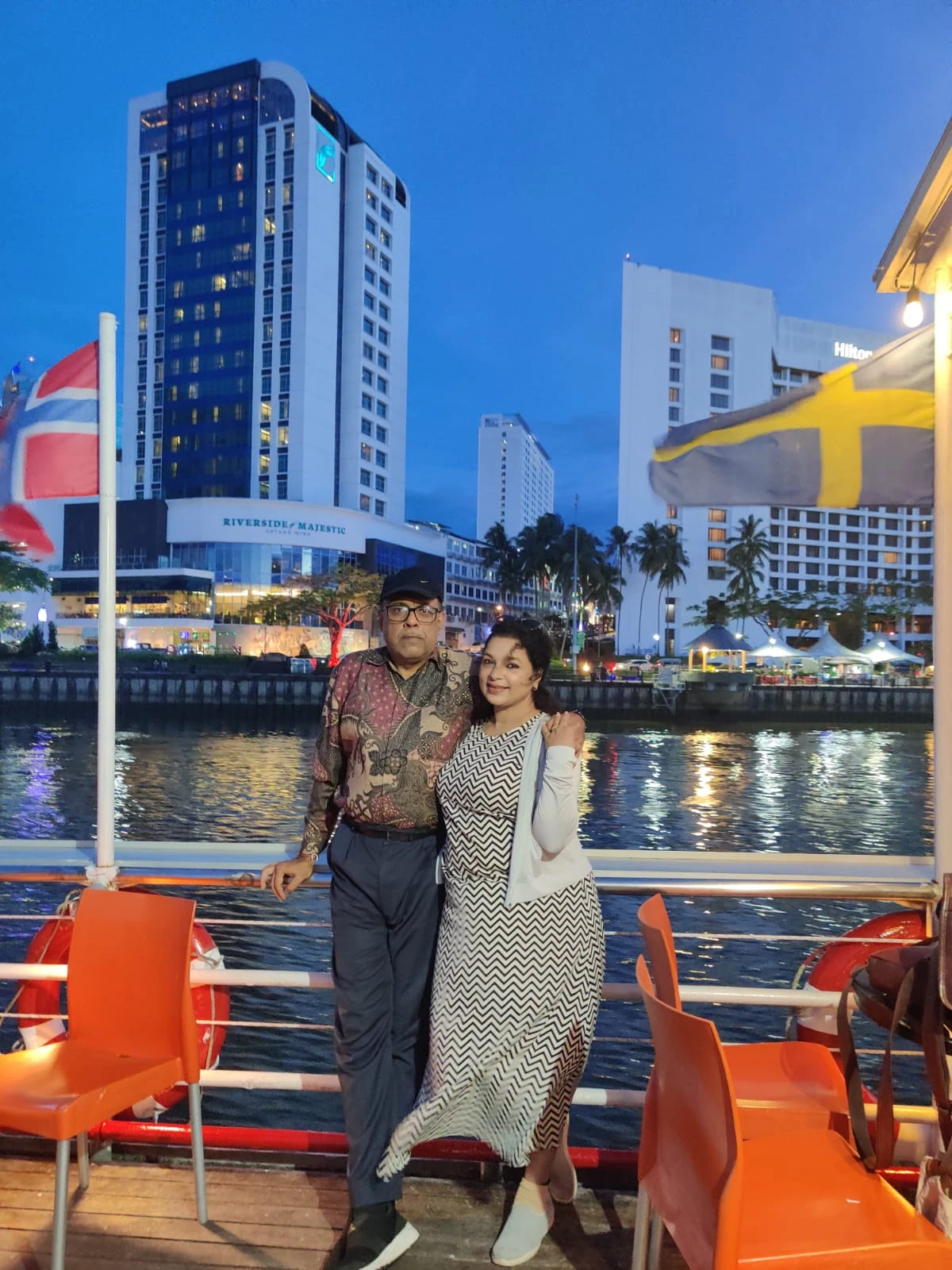 Couple travelers standing on the open deck of the Sarawak River cruise boat at dusk with Kuching waterfront in the background