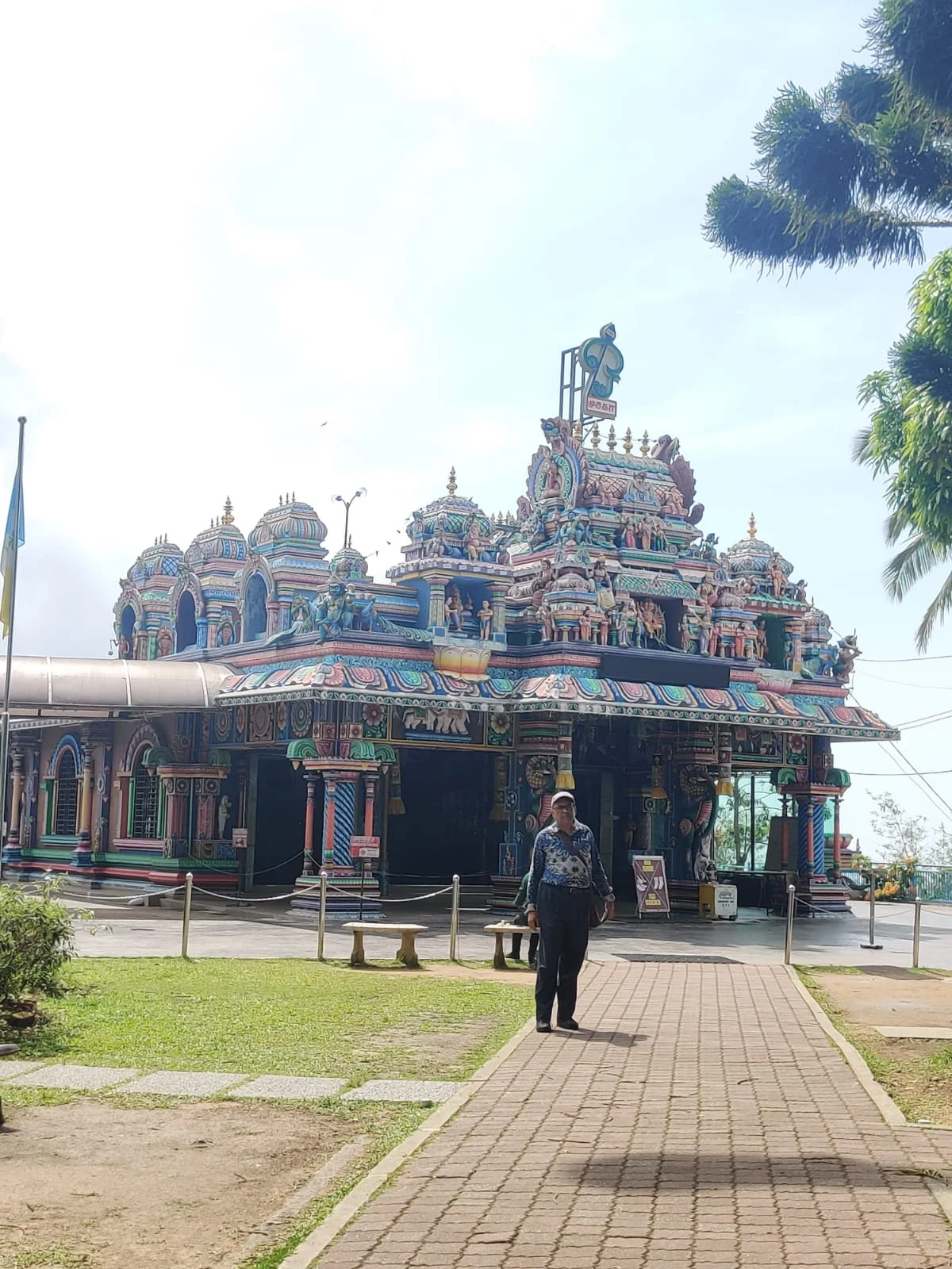 Male traveler standing before the colorful Dravidian facade and Vel emblem of Sri Aruloli Thirumurugan Temple on Penang Hill