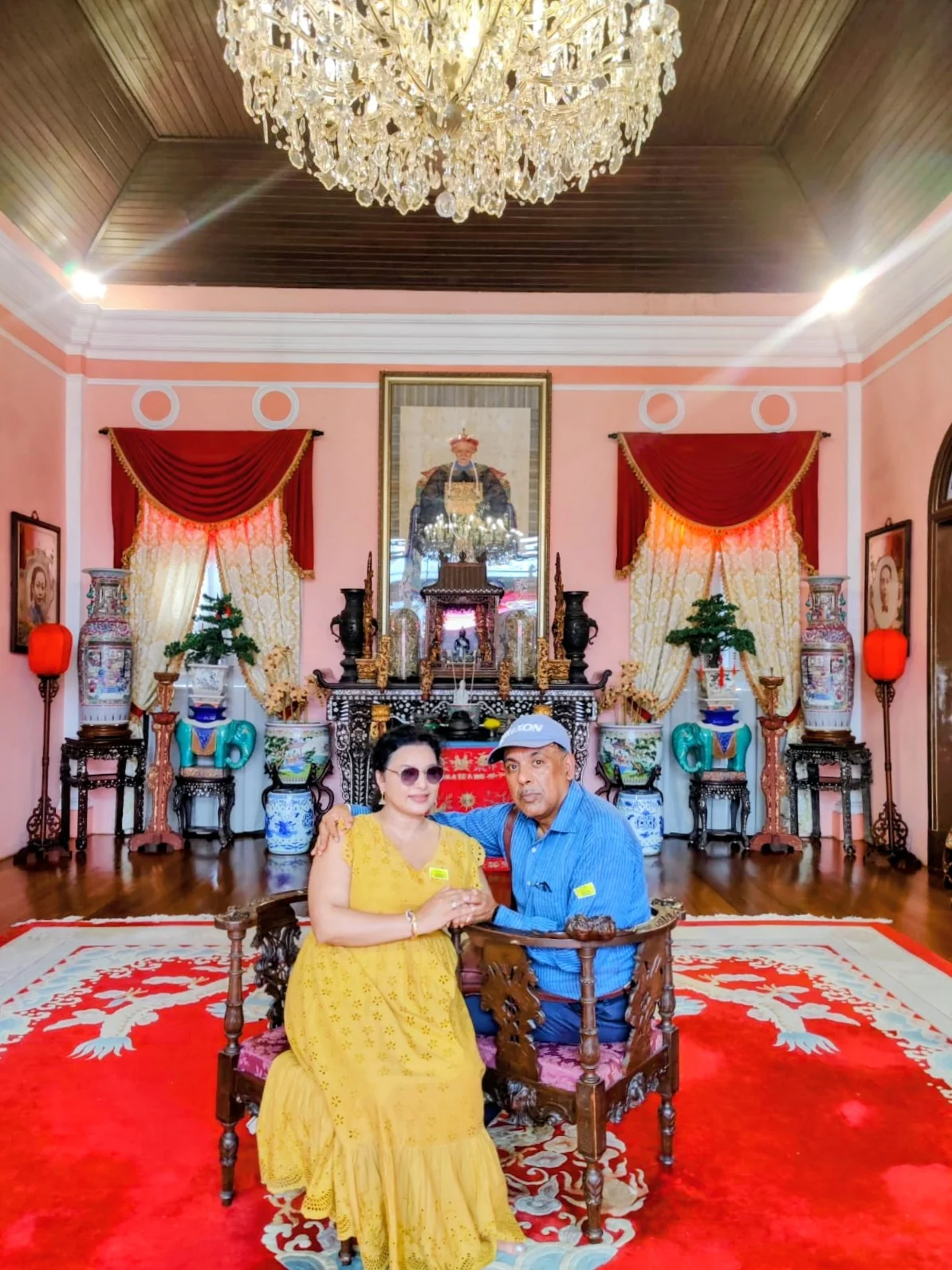 Couple travelers seated in the grand ancestral hall of the Chinese heritage home beneath crystal chandeliers and gold-leaf screens