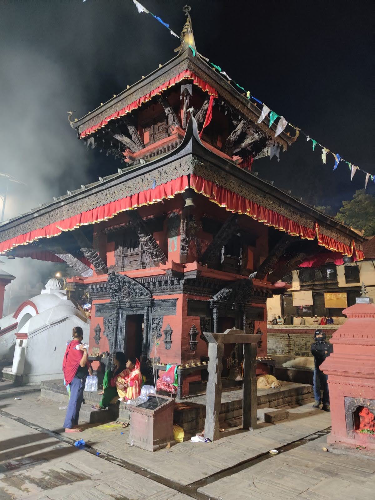Illuminated multi-tiered pagoda shrine with red frills and intricate carved wood struts in the Pashupatinath courtyard