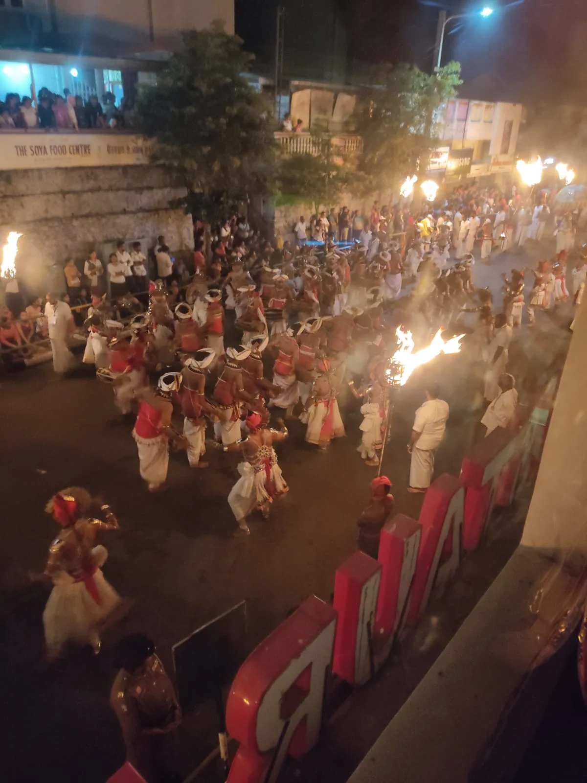 High angle perspective from the restaurant balcony showing hundreds of torch-bearing dancers and drummers marching through crowded Kandy streets