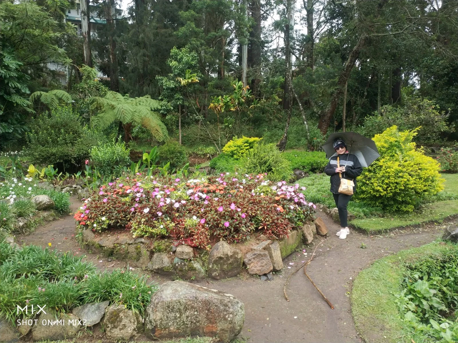 Traveler holding an umbrella in Victoria Park surrounded by vibrant stone-bordered flowerbeds and lush greenery