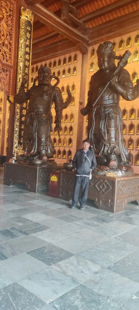 Male traveler standing beside colossal bronze Dharmapala guardian statues in the grand hall of Bai Dinh Pagoda