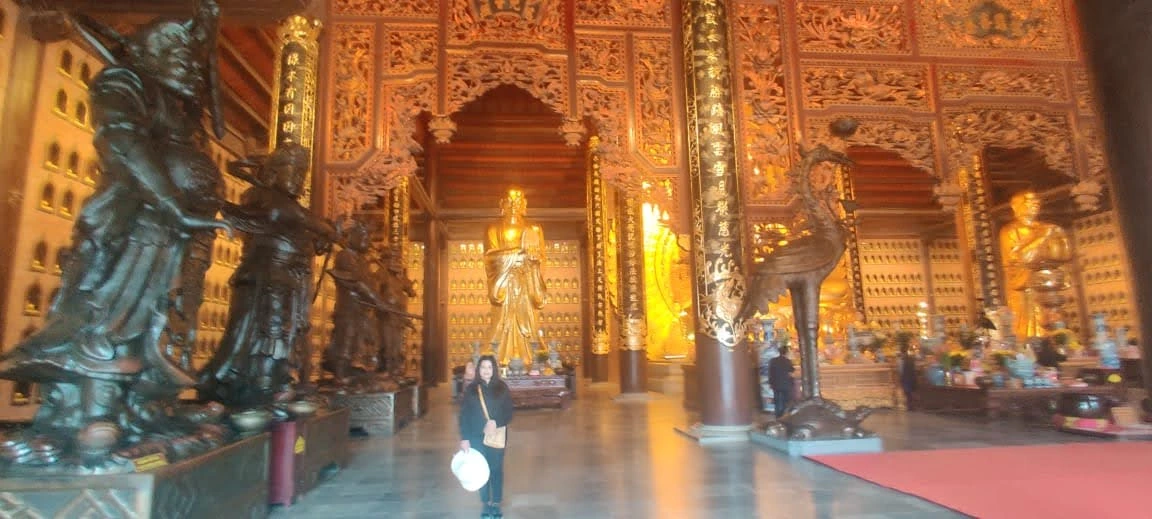 Panoramic view inside the grand sanctuary hall of Bai Dinh Pagoda with bronze guardian deities and standing Golden Buddha