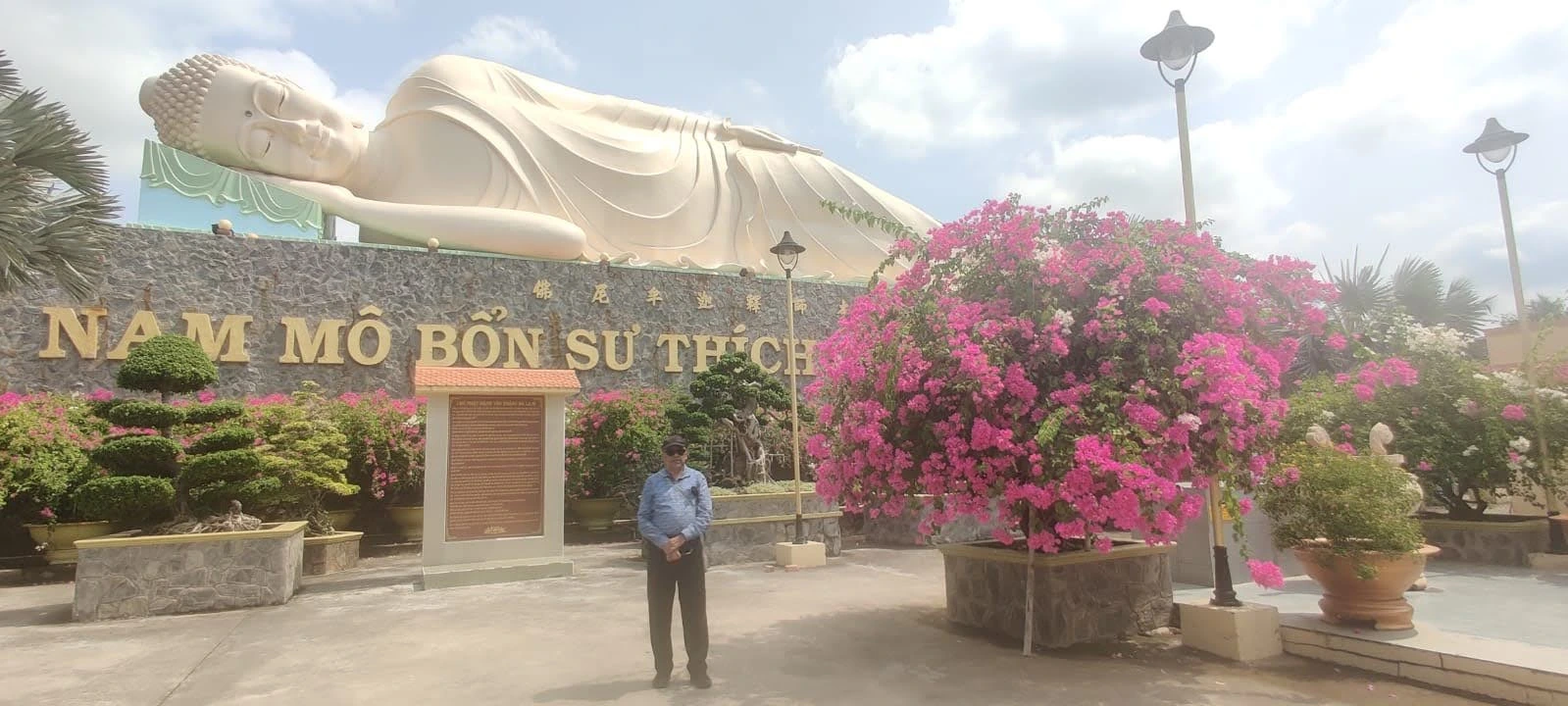 Male traveler standing before the colossal Reclining Buddha at Vinh Trang Pagoda surrounded by vibrant pink bougainvillea