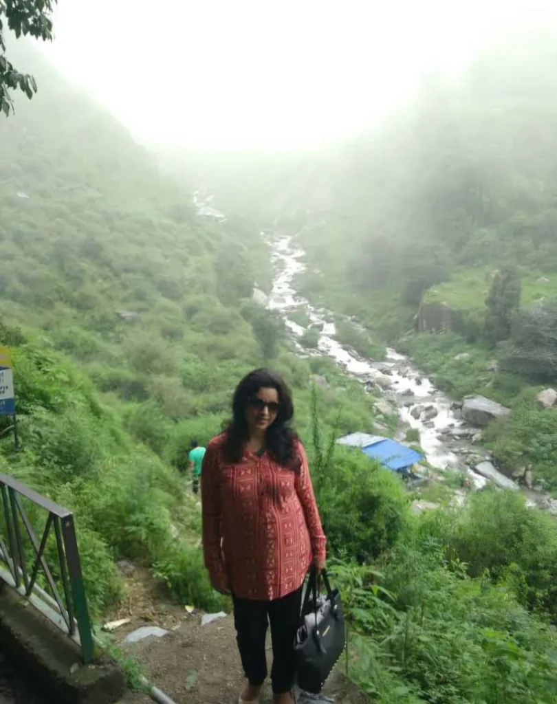 Traveler standing near a path with the cascading Bhagsunath Waterfall in the misty valley behind her